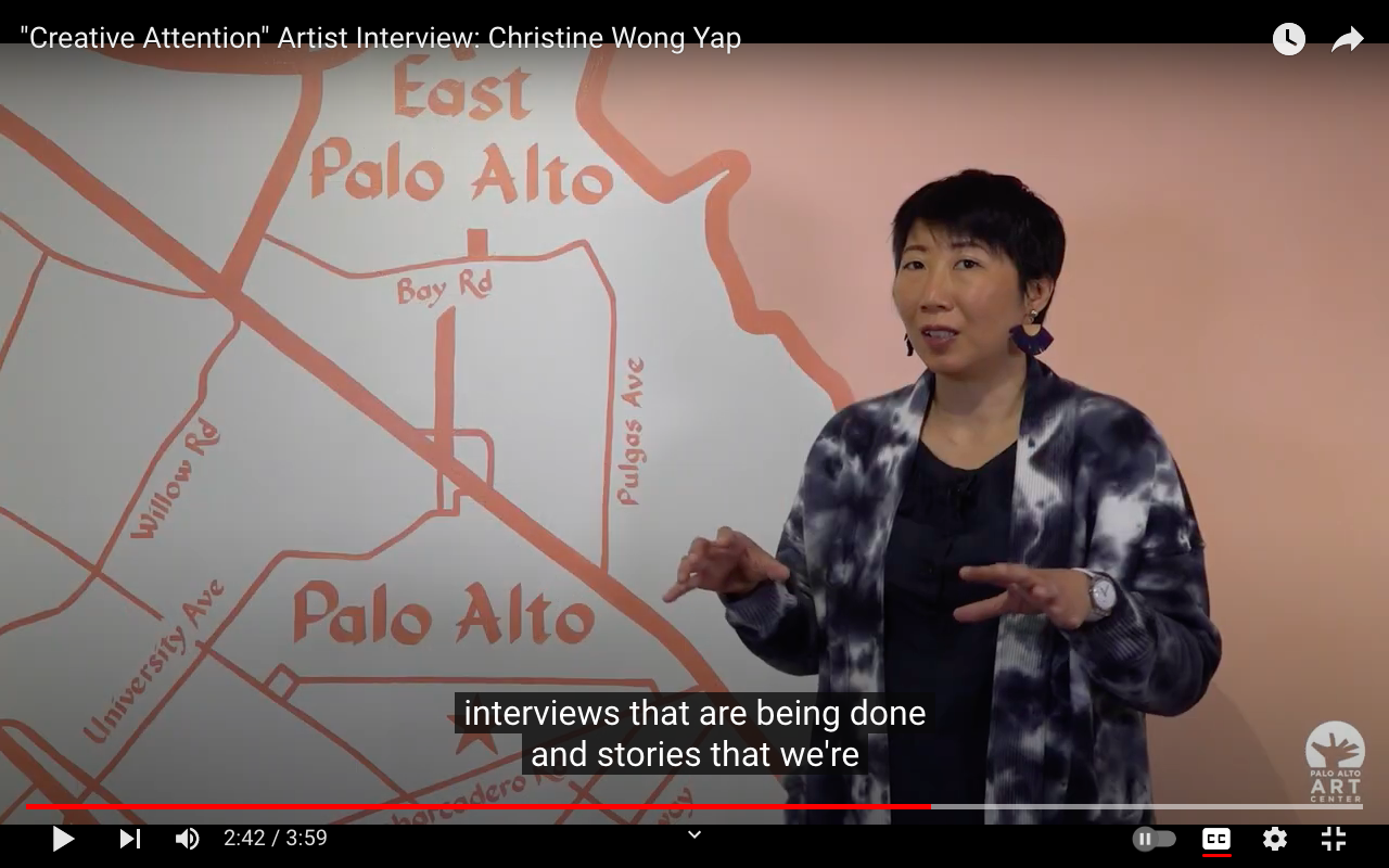 still from video of artist speaking in front of a map of palo alto and east palo alto painted on the wall